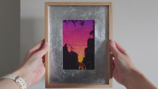 Painting on canvas in a silver and wood frame. Painting shows a purple to pink to yellow gradient sunset that silhouettes a cityscape, on the rooftops are two gargoyles. Held in a pair of hands to show the paintings details. 
